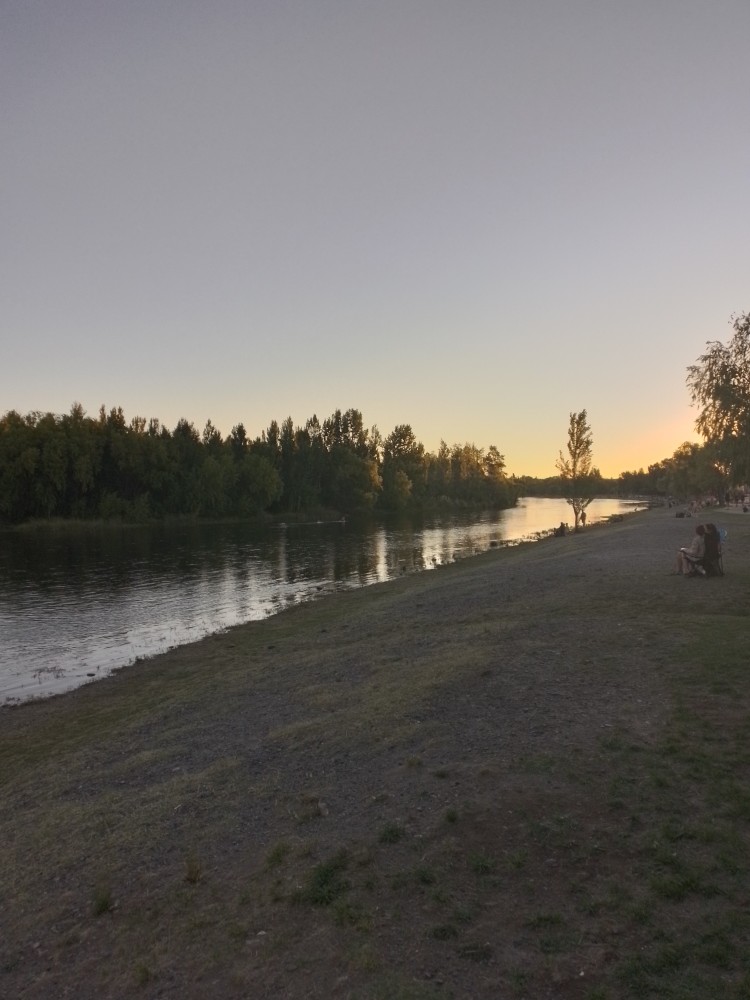 A photograph of a beautiful riverside afternoon. The sun has set, and its golden crown shines in the sky uncontested by clouds or stars. There's still a few people sitting and chatting.