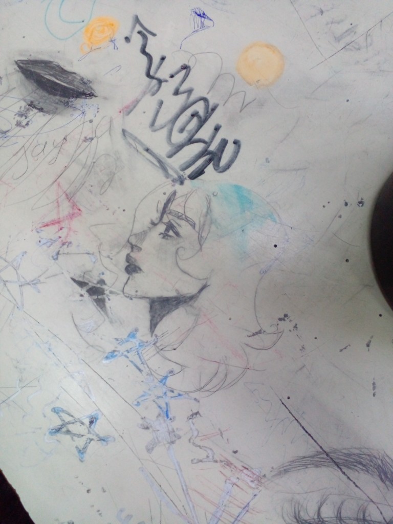 A beautiful anime youth with long hair drawn on a school table, surrounded by other colorful doodles.
