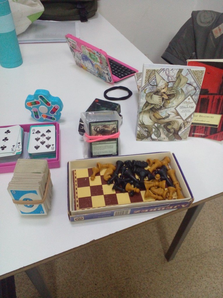 An assortment of items on a school table. There's a portable chess, a few books and manga, a fidget toy puzzle with colored balls, playing cards, Uno cards and Magic: The Gathering cards. When we're at the end of the year I bring these for fun.