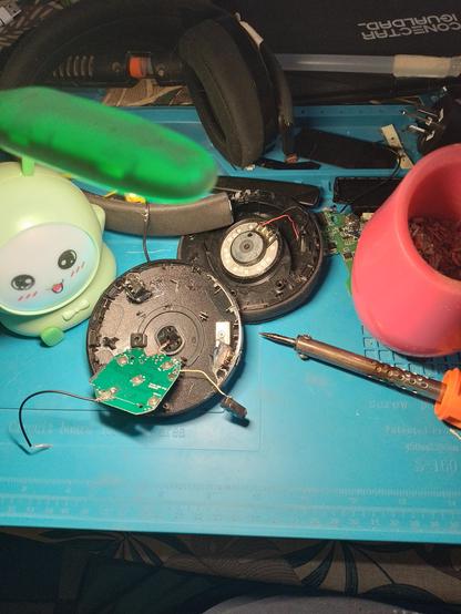 A cyan rubber mat littered with electronics, pieces of a headset, a green creature lamp, and a pink plastic mate.
