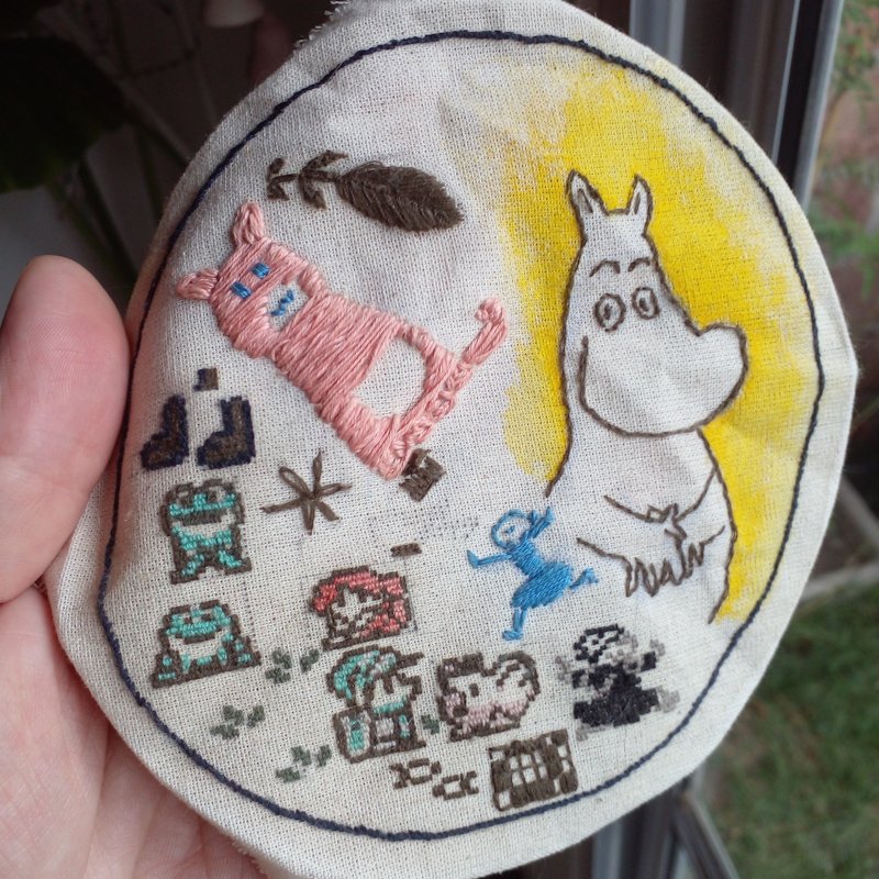 A round patch of linen with many characters embroidered in a very fine pixel art style. There's Link, Marin, a chicken and two frogs from The Legend of Zelda: Link's Awakening, musical notes and fish, leaves, two Falins hopping from a flashback in DunMeshi, a pink cat with a silly face, and Moomin with his hands on his tummy.