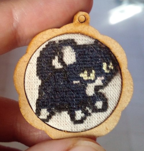 A round keychain with linen on the center. Embroidered on it is a tuxedo cat done in a pixel art style.