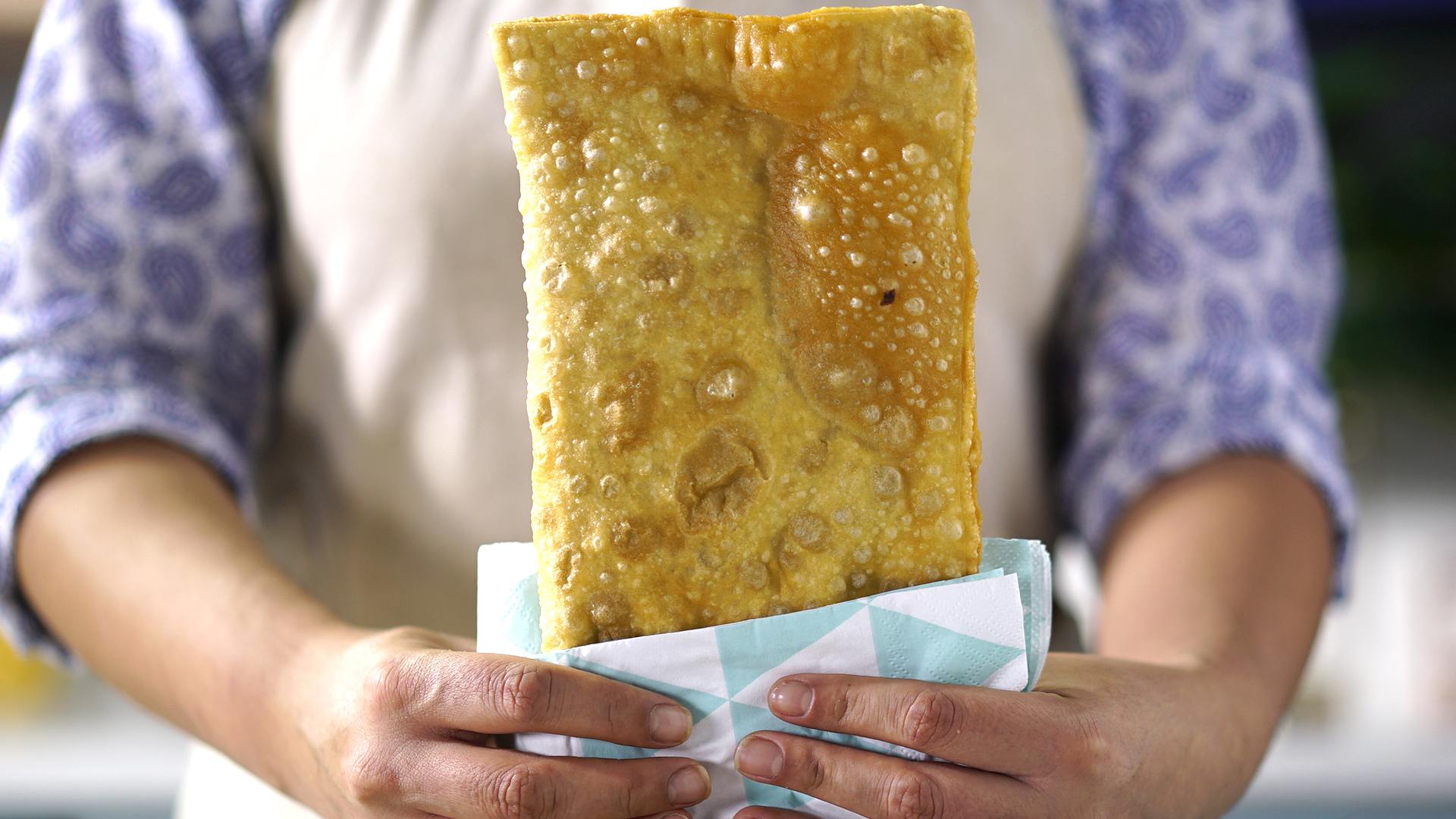 A fried bread a bit smaller than an A4 sheet of paper, held by a woman in cooking attire.